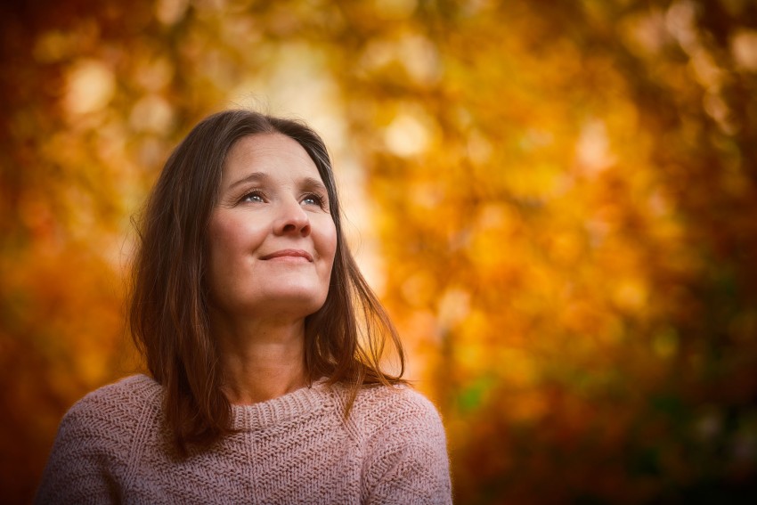 Kvinne med brunt hår og rosa genser, smilende og ser opp, mot en bakgrunn av oransje høstløvverk. Foto: Nathan Lediard, Lediard Foto AS.