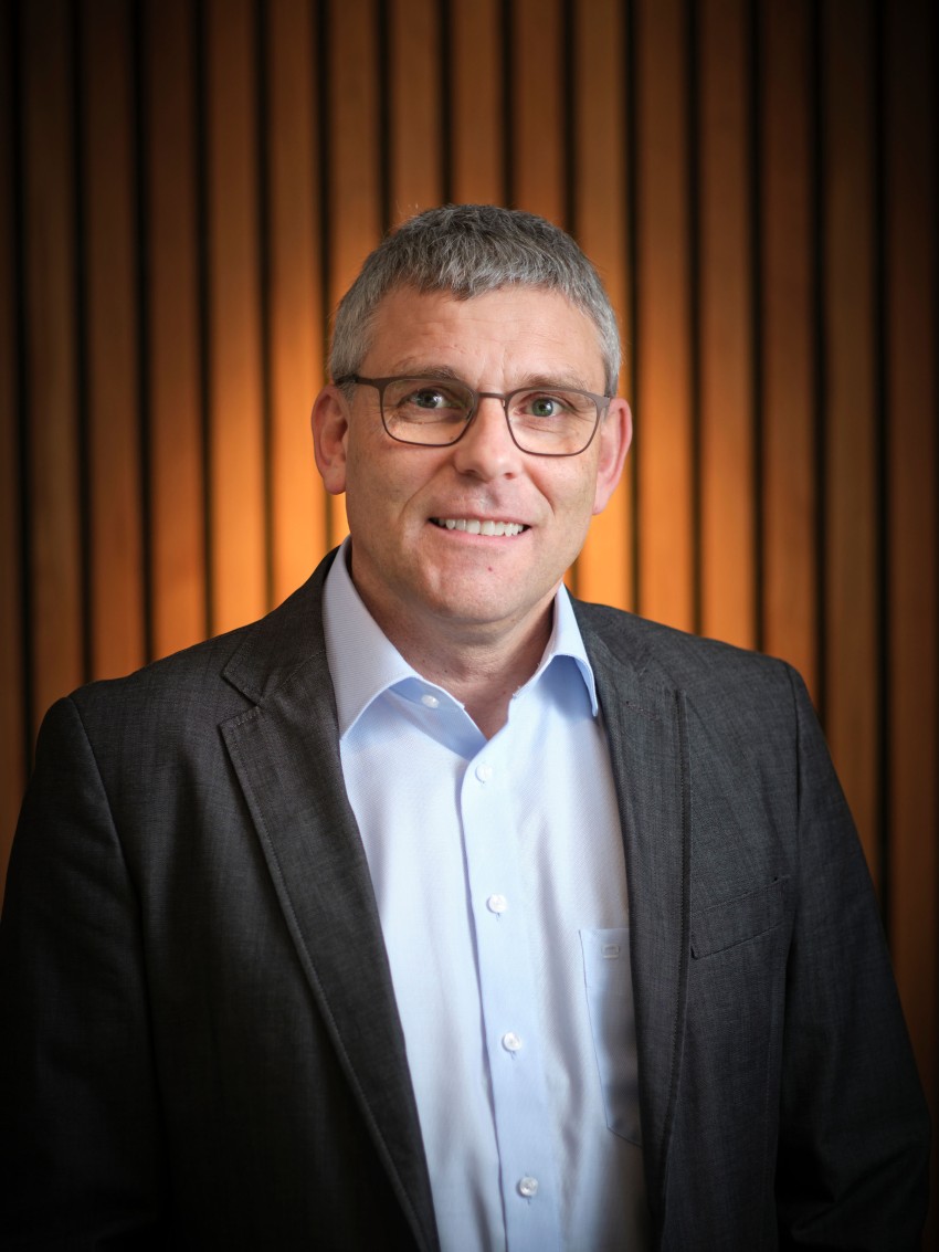 Mann i briller og dress står foran en trepanelvegg og smiler til kameraet. Foto: Nathan Lediard, Lediard Foto AS.