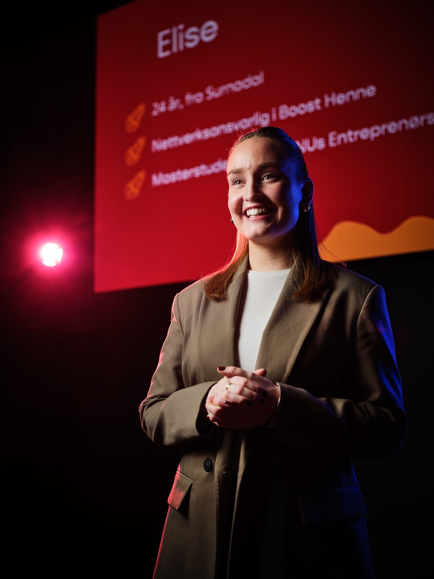 Smilende kvinne i blazer står på scenen med et presentasjonslysbilde bak seg. Foto: Nathan Lediard, Lediard Foto AS.