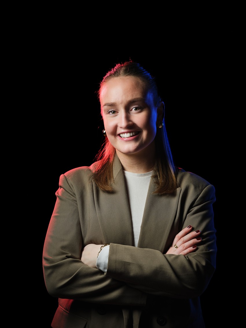 Smilende person med korslagte armer iført en brun blazer, med rødt og blått lys mot en svart bakgrunn. Foto: Nathan Lediard, Lediard Foto AS.