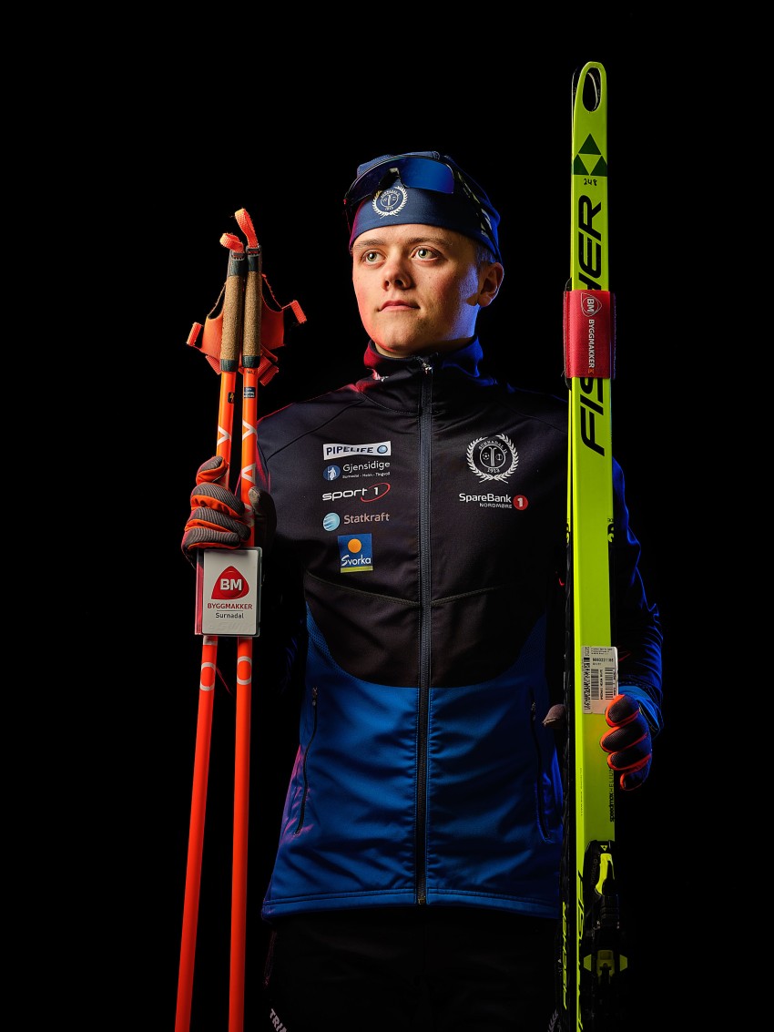 En skiløper i vinterutstyr holder røde skistaver og en grønn ski, stående mot en mørk bakgrunn. Foto: Nathan Lediard, Lediard Foto AS.