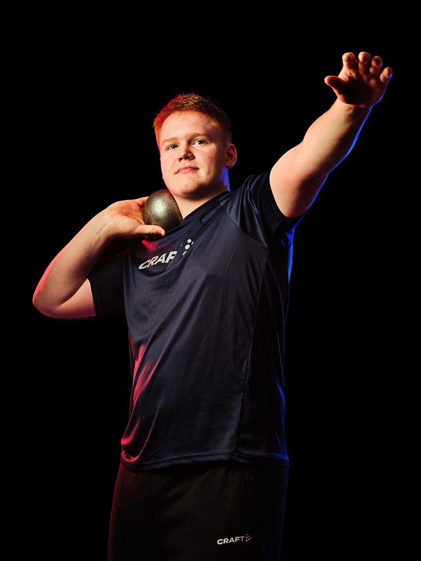 Idrettsutøver i mørk skjorte klar til å kaste et kulestøt, armen utstrakt, på mørk bakgrunn. Foto: Nathan Lediard, Lediard Foto AS.