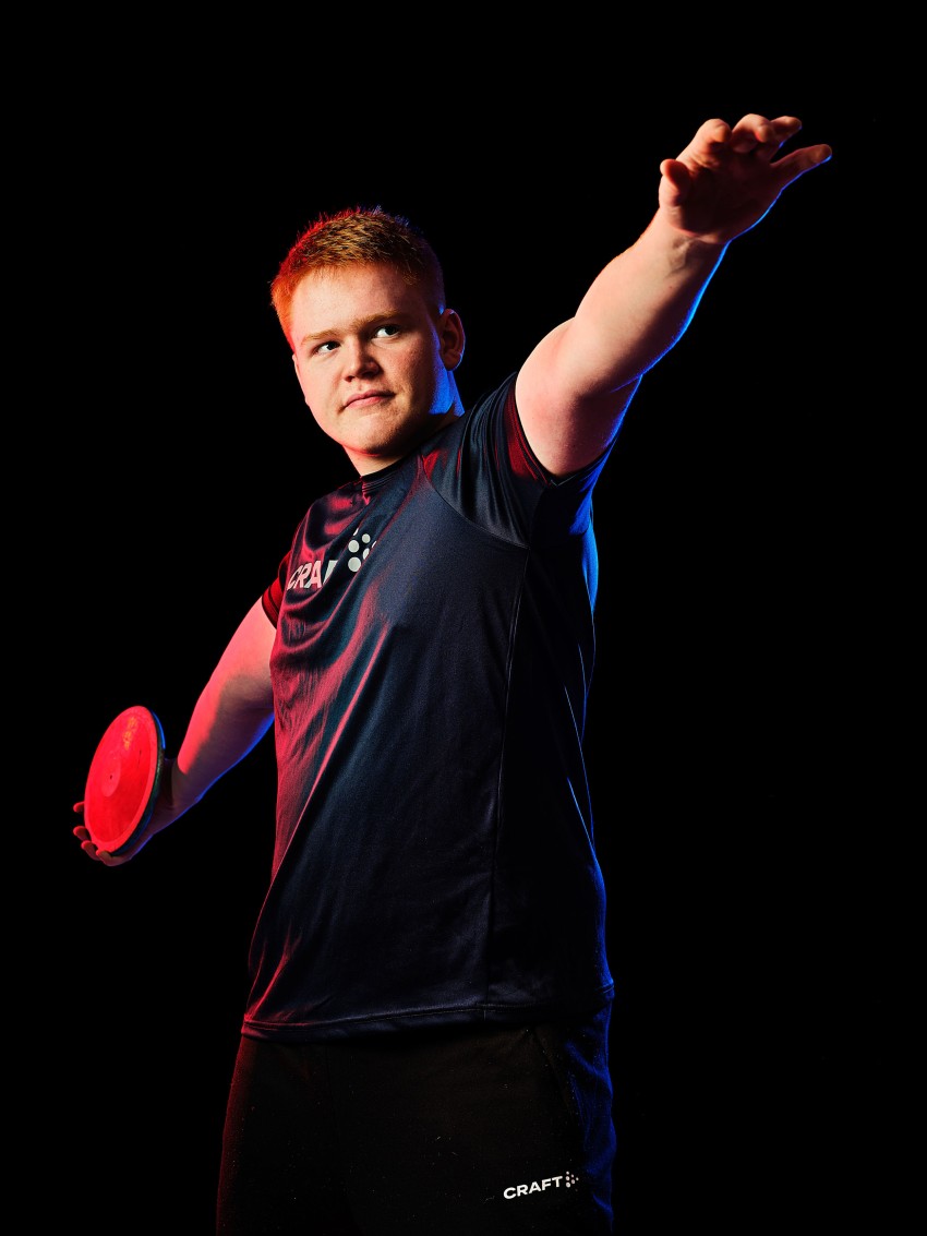 En idrettsutøver i mørkt sportsantrekk poserer med en rød diskos mot en svart bakgrunn, opplyst av fargede lys. Foto: Nathan Lediard, Lediard Foto AS.