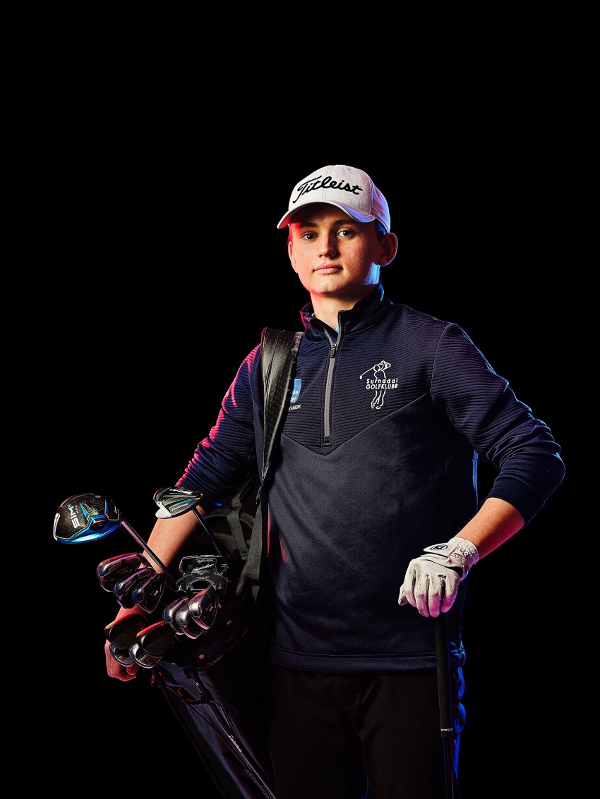 Golfspiller som holder en pose med køller, iført hette og hanske, står selvsikkert mot en svart bakgrunn. Foto: Nathan Lediard, Lediard Foto AS.
