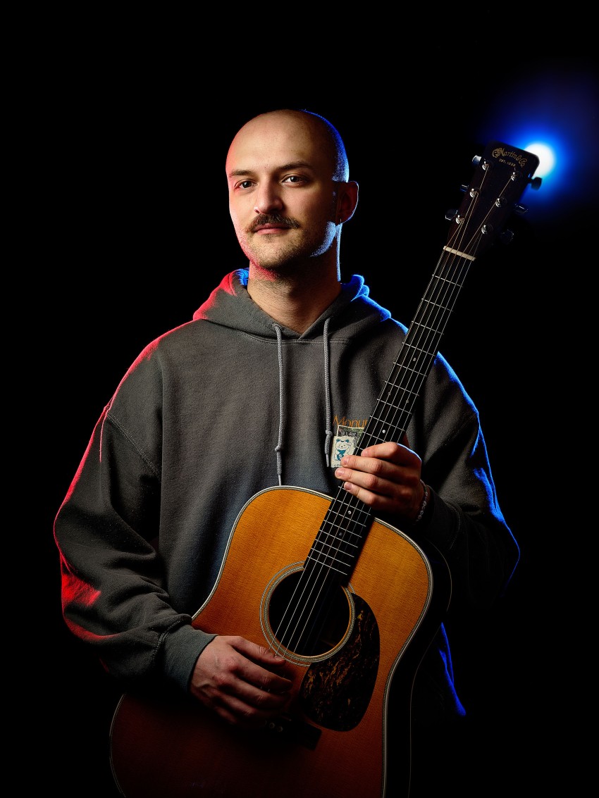 Person i en hettegenser som holder en akustisk gitar mot en mørk bakgrunn med blått og rødt lys. Foto: Nathan Lediard, Lediard Foto AS.