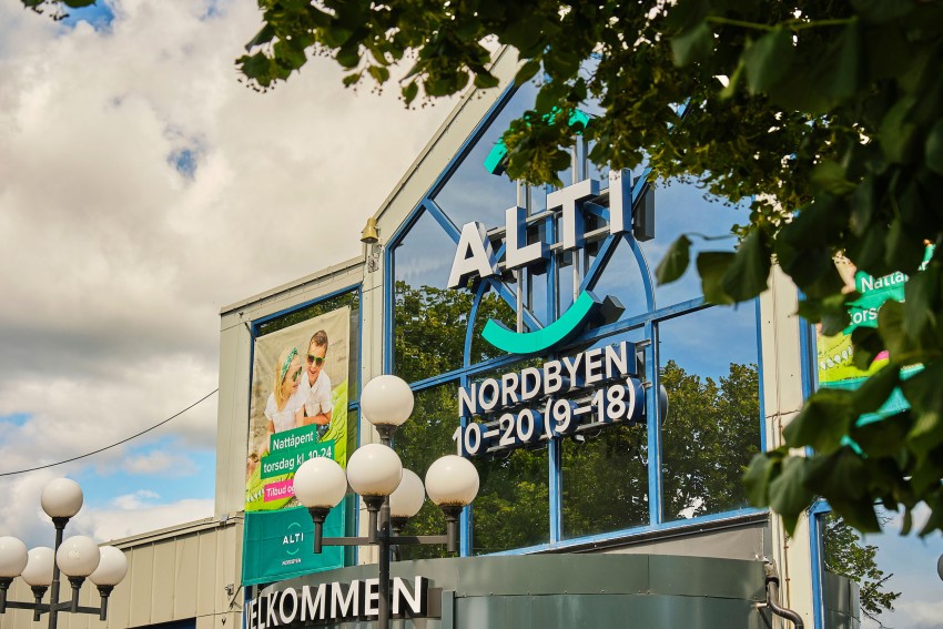 Kjøpesenterinngang med "ALTI" og "NORDBYEN" skilt, trær og gatelykter. Foto: Nathan Lediard, Lediard Foto AS.