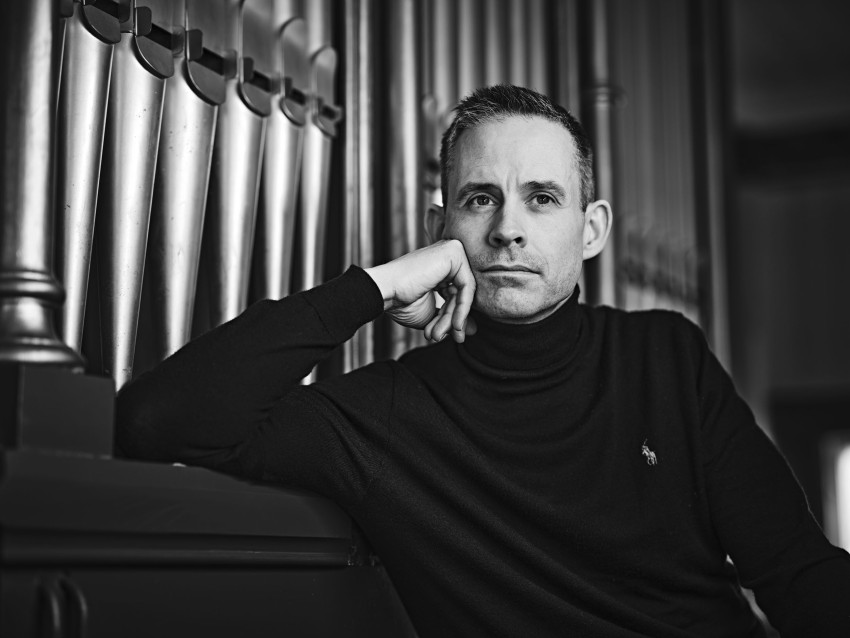 Mann i svart turtleneck, hvilende hake på hånden, poserer ved store orgelpiper i et svart-hvitt bilde. Foto: Nathan Lediard, Lediard Foto AS.