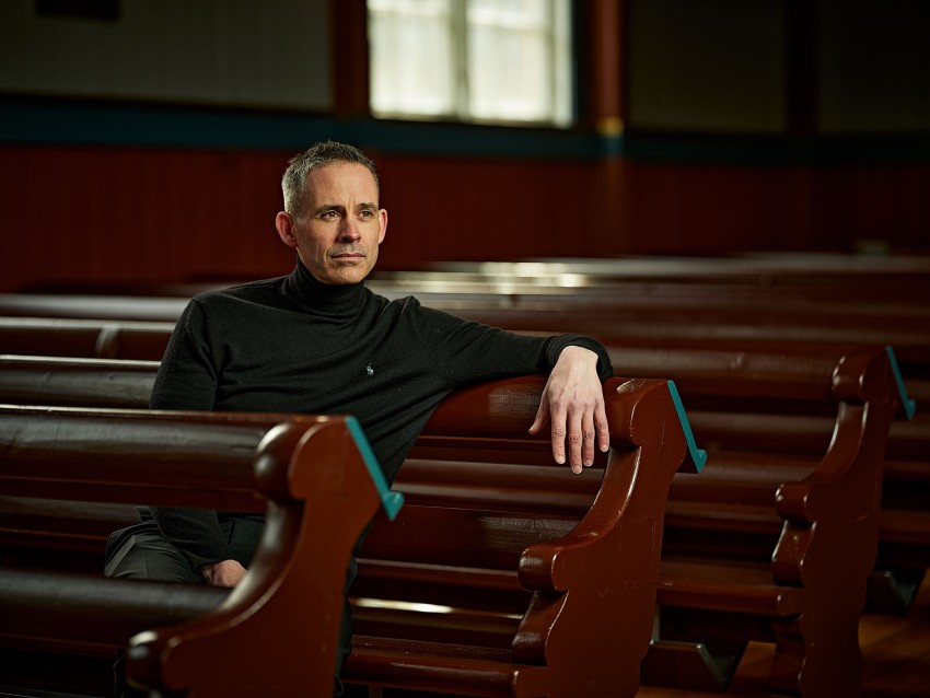 Mann som sitter på kirkebenker i tre, iført en svart turtleneck, hviler armen på ryggstøtten og ser ettertenksom ut. Foto: Nathan Lediard, Lediard Foto AS.