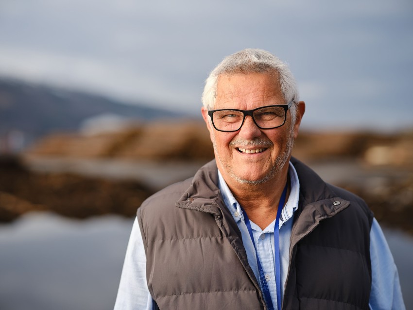 Smilende eldre mann med briller og grå vest står utendørs med et uskarpt naturlandskap i bakgrunnen. Foto: Nathan Lediard, Lediard Foto AS.