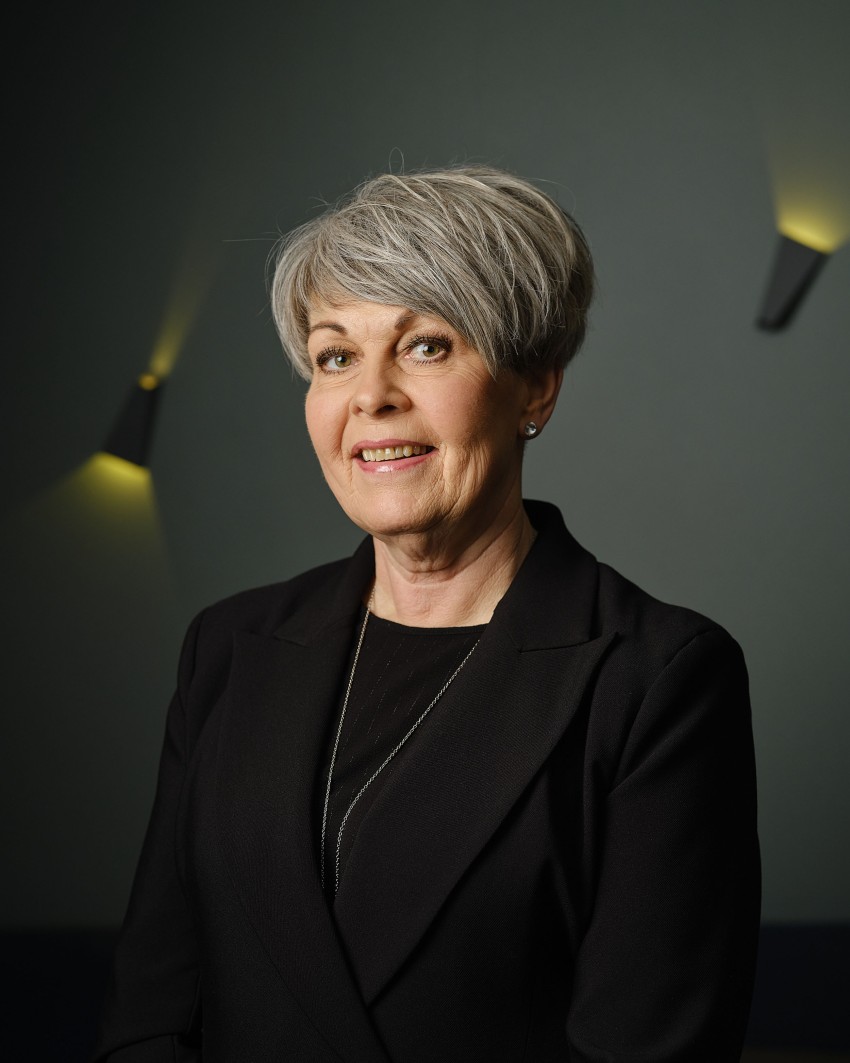 Person med kort grått hår og en svart blazer smilende, stående mot en mørk bakgrunn med varme lys. Foto: Nathan Lediard, Lediard Foto AS.