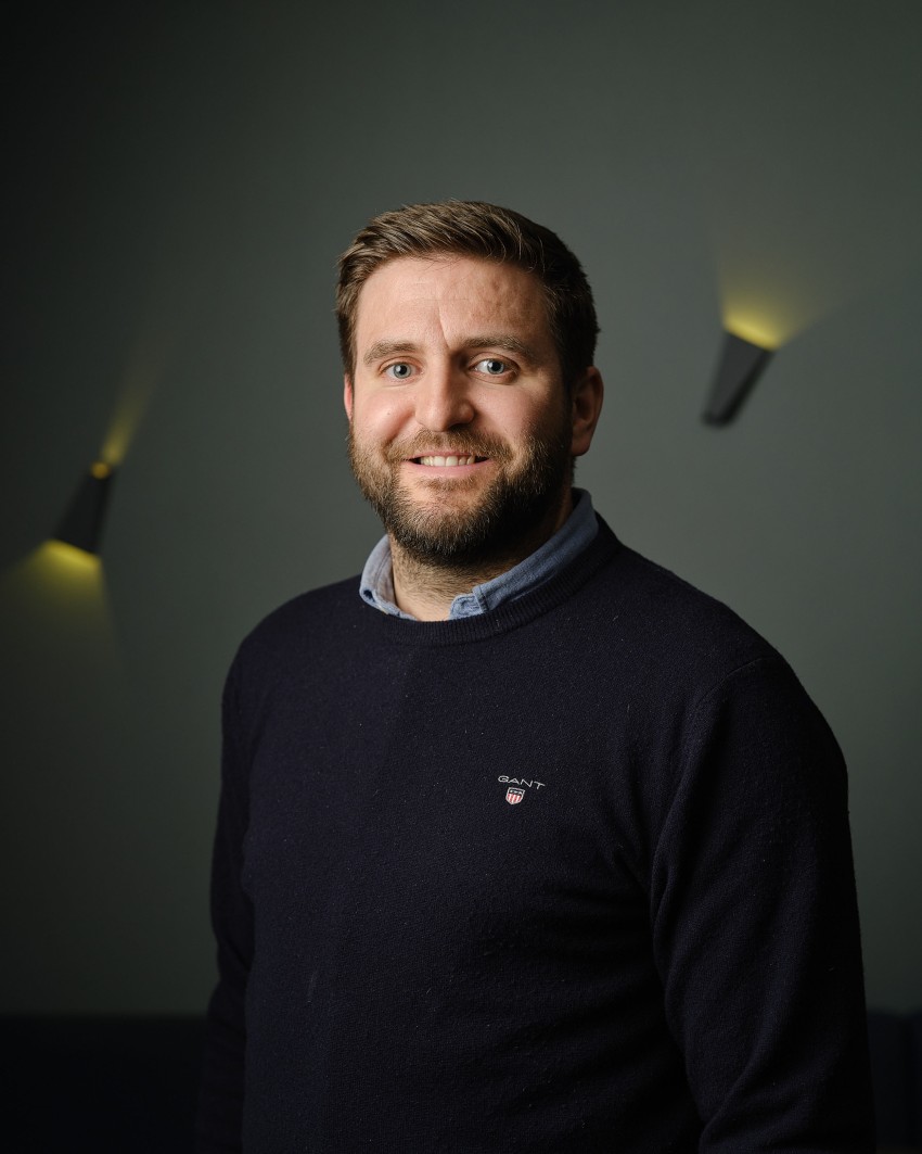 Smilende mann med skjegg i mørk genser står mot en mørk vegg med lys. Foto: Nathan Lediard, Lediard Foto AS.