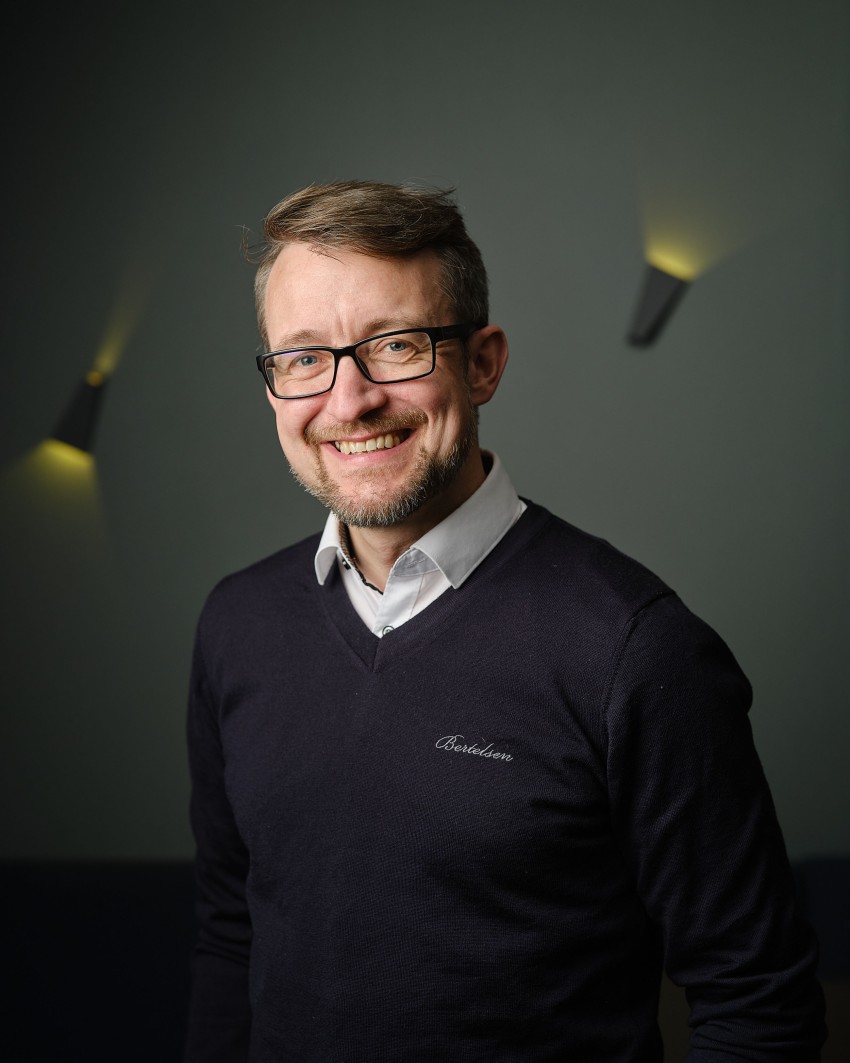Mann med briller og skjegg smilende, iført en svart genser over en hvit skjorte, stående mot en mørk bakgrunn. Foto: Nathan Lediard, Lediard Foto AS.
