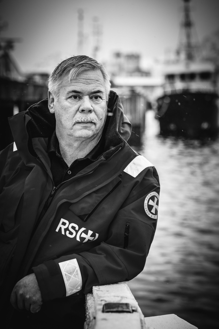 Mann i mørk jakke lener seg på et rekkverk nær vann, med skip uskarpt i bakgrunnen. Svart-hvitt bilde. Foto: Nathan Lediard, Lediard Foto AS.