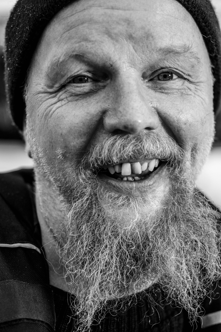Skjeggete mann i lue som smiler varmt, svart-hvitt nærbildeportrett. Foto: Nathan Lediard, Lediard Foto AS.