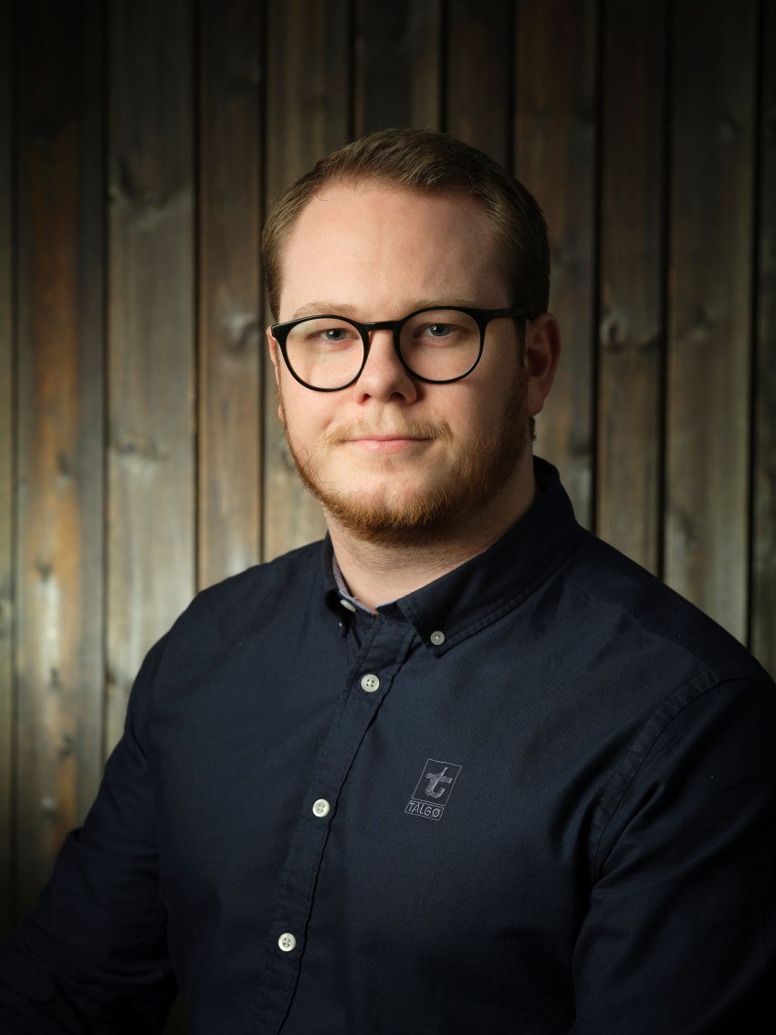 Mann med briller og skjegg iført en mørk skjorte, stående mot et trebakgrunn. Foto: Nathan Lediard, Lediard Foto AS.