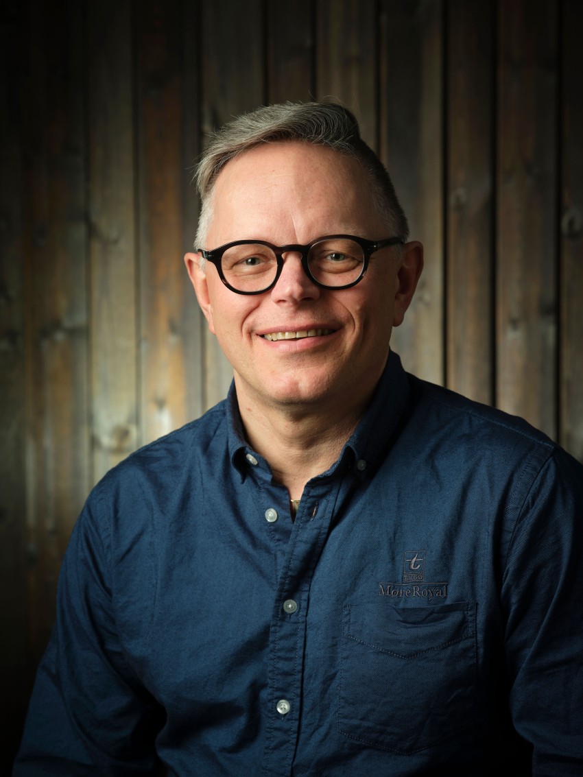 Smilende person med briller og blå skjorte, sittende mot en trebakgrunn. Foto: Nathan Lediard, Lediard Foto AS.