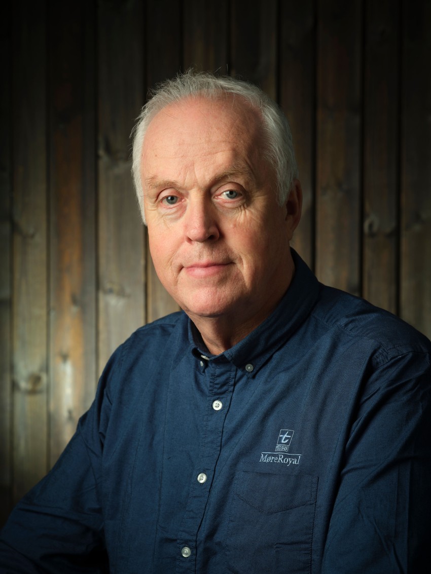 Eldre mann i blå skjorte sitter foran en trebakgrunn og ser på kameraet. Foto: Nathan Lediard, Lediard Foto AS.