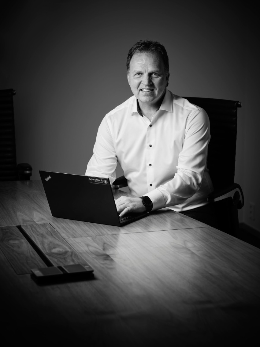 Mann i hvit skjorte sitter ved et bord med en bærbar datamaskin og smiler i et svakt opplyst rom. Foto: Nathan Lediard, Lediard Foto AS.