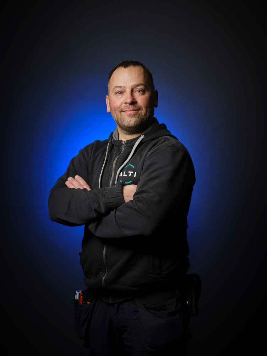 Mann i mørk hettegenser med armene i kors, smilende foran en blå halo-bakgrunn. Foto: Nathan Lediard, Lediard Foto AS.