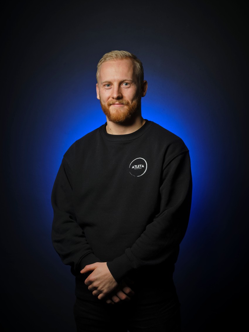 En skjeggete mann i svart genser står mot en blåopplyst mørk bakgrunn og smiler lett med hendene sammen. Foto: Nathan Lediard, Lediard Foto AS.