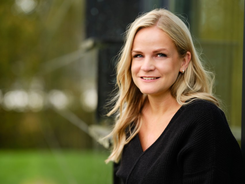 Smilende kvinne med langt blondt hår i en svart topp, som sitter utendørs med en grønn uskarp bakgrunn. Foto: Nathan Lediard, Lediard Foto AS.