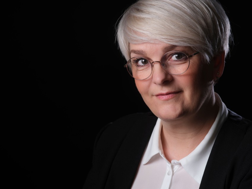 Person med kort hvitt hår og briller, iført svart jakke og hvit skjorte, mot mørk bakgrunn. Foto: Nathan Lediard, Lediard Foto AS.