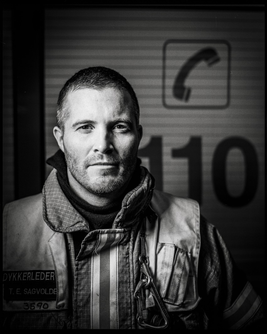 Svart-hvitt-bilde av en brannmann i uniform som står foran et nødtelefonskilt. Foto: Nathan Lediard, Lediard Foto AS.