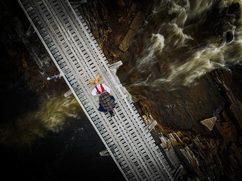 Person i et rødt skjørt liggende på en trebro over en brusende elv, omgitt av steiner, sett ovenfra. Foto: Nathan Lediard, Lediard Foto AS.