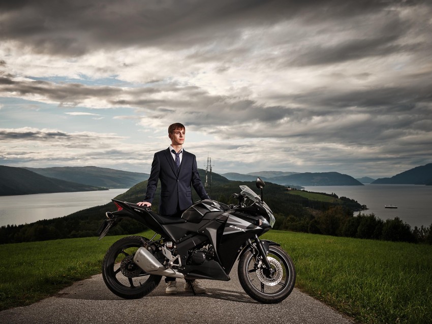 Mann i dress står ved siden av en svart motorsykkel på en høyde, med en vakker innsjø og overskyet himmel i bakgrunnen. Foto: Nathan Lediard, Lediard Foto AS.