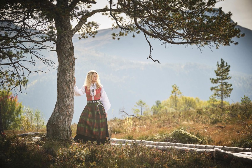 Kvinne i tradisjonell kjole står ved siden av et tre i et naturskjønt fjellandskap. Foto: Nathan Lediard, Lediard Foto AS.