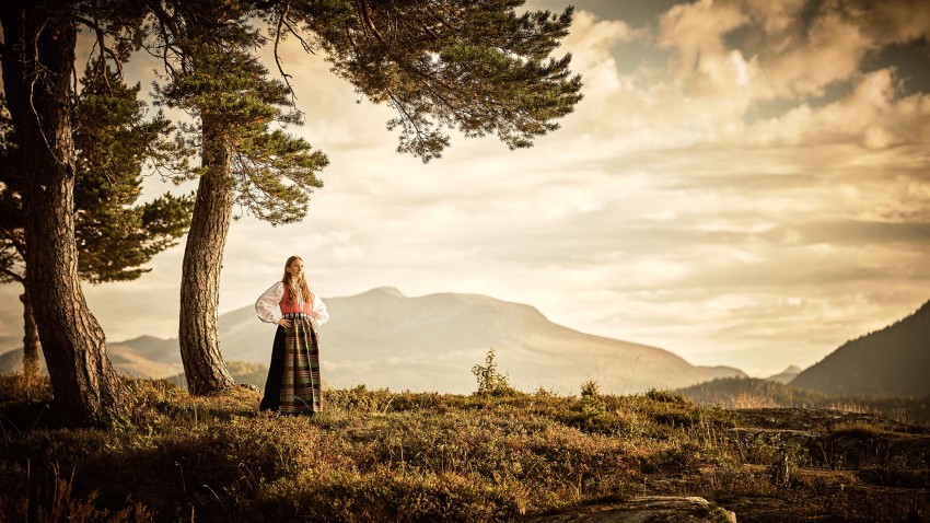 Kvinne i tradisjonell kjole står blant trær, med utsikt over fjellene under en overskyet himmel. Foto: Nathan Lediard, Lediard Foto AS.