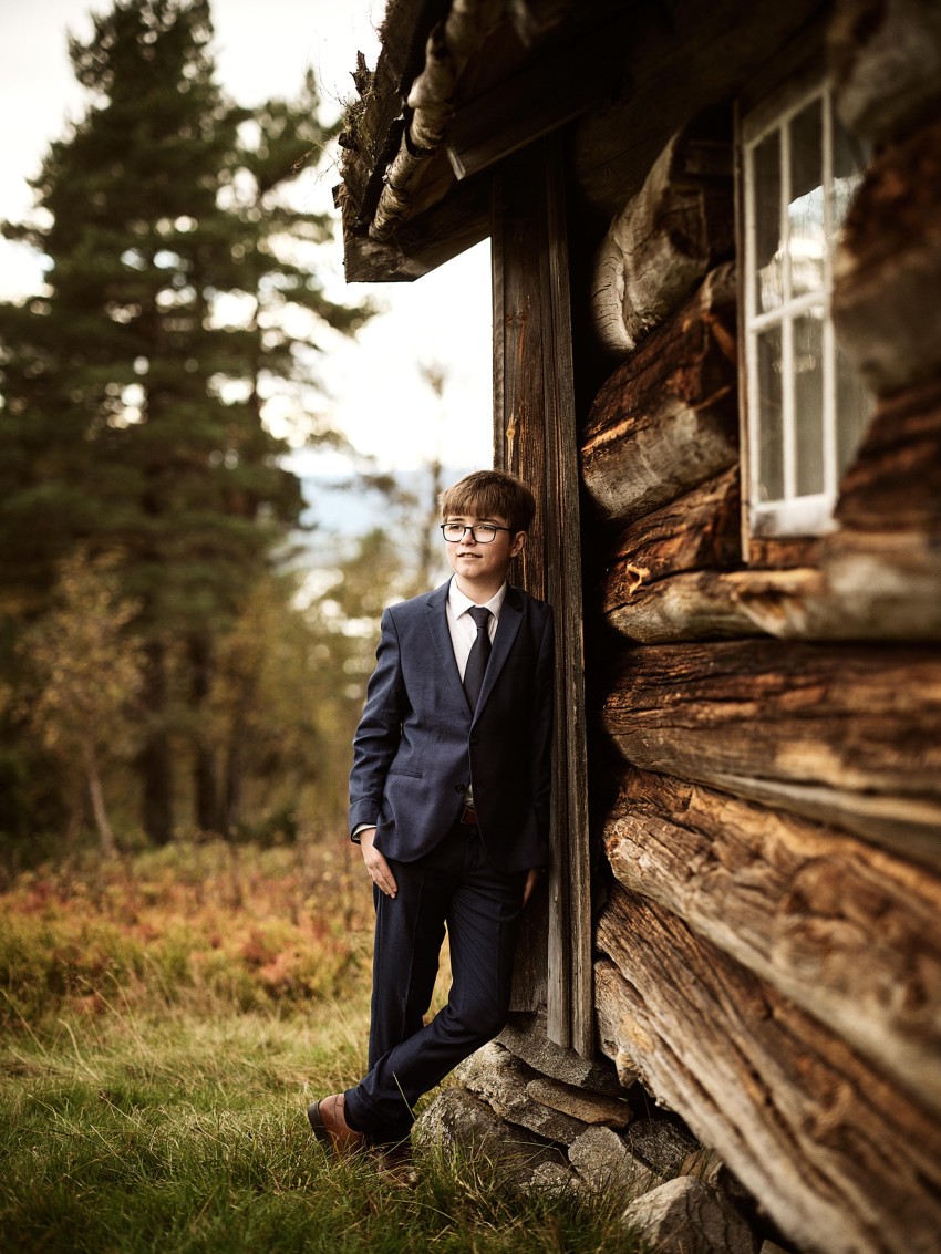 Gutt i blå dress lener seg mot en rustikk trehytte, omgitt av trær og gress. Foto: Nathan Lediard, Lediard Foto AS.