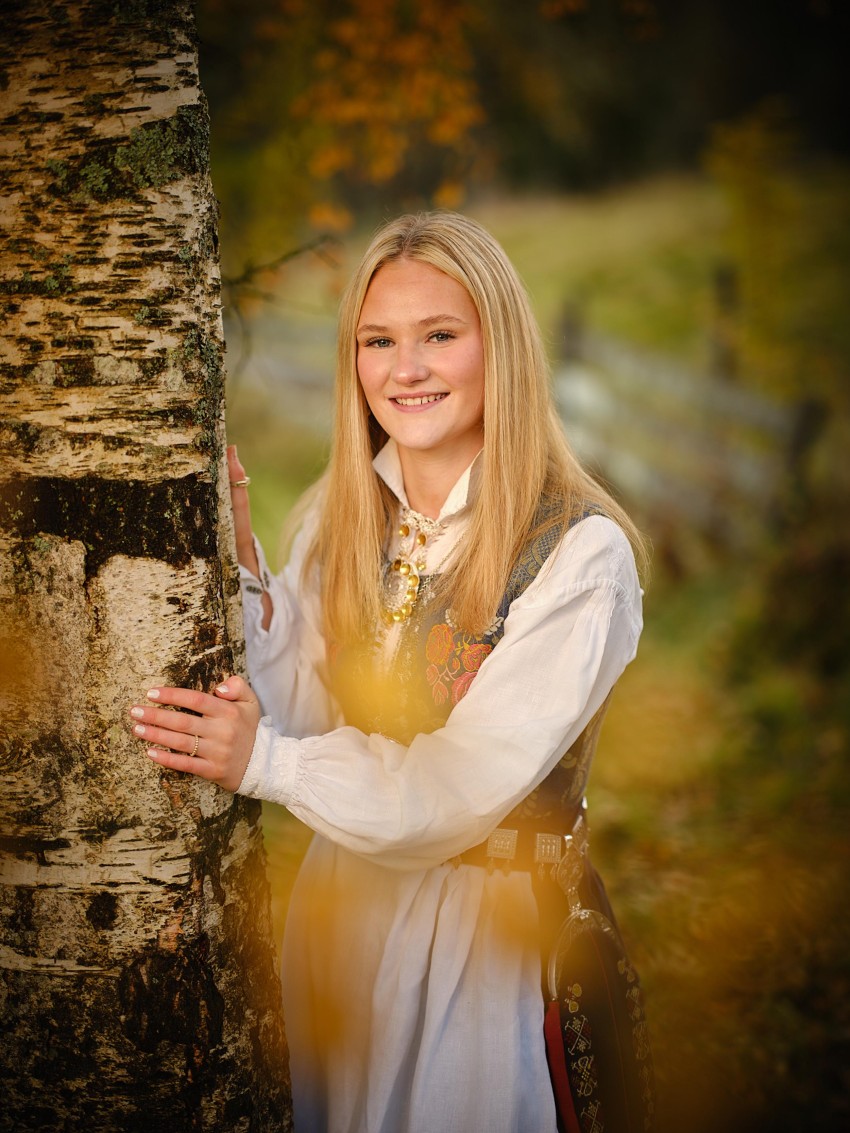 Smilende jente i tradisjonell kjole står ved et tre i grønne, utendørs omgivelser. Foto: Nathan Lediard, Lediard Foto AS.