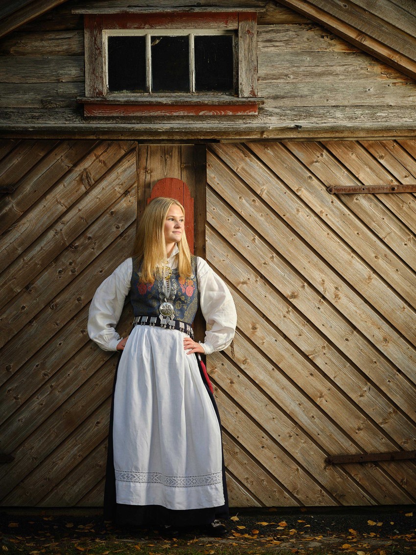 Ung kvinne i tradisjonell kjole står mot en låvedør av tre, hendene på hoftene og ser til siden. Foto: Nathan Lediard, Lediard Foto AS.