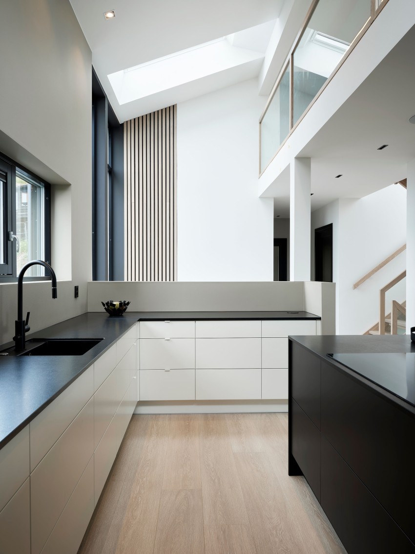 Moderne kjøkken med sorte benkeplater, hvite skap, store vinduer, og høyt under taket med takvindu. Foto: Nathan Lediard, Lediard Foto AS.