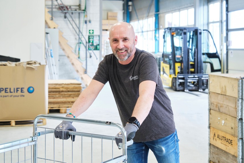 Smilende mann med hansker står ved en metallvogn i et lager med bokser og en gaffeltruck i bakgrunnen. Foto: Nathan Lediard, Lediard Foto AS.