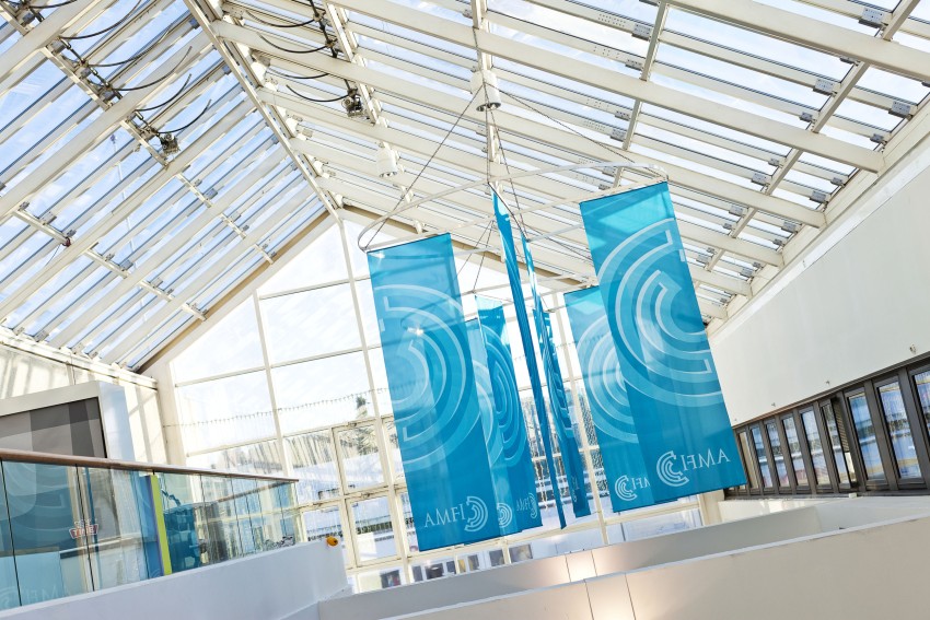 Lyst atrium med store blå bannere som viser hvite logoer og mønstre hengende fra glasstaket. Foto: Nathan Lediard, Lediard Foto AS.