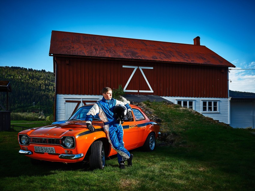 Racer i blå dress lener seg på en oransje veteranbil foran en rød låve, med en svart hjelm. Foto: Nathan Lediard, Lediard Foto AS.