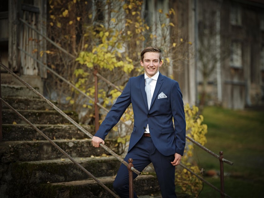 Ung mann i blå dress står på utendørstrinn og holder rekkverket. En rustikk bygning og høsttrær er i bakgrunnen. Foto: Nathan Lediard, Lediard Foto AS.