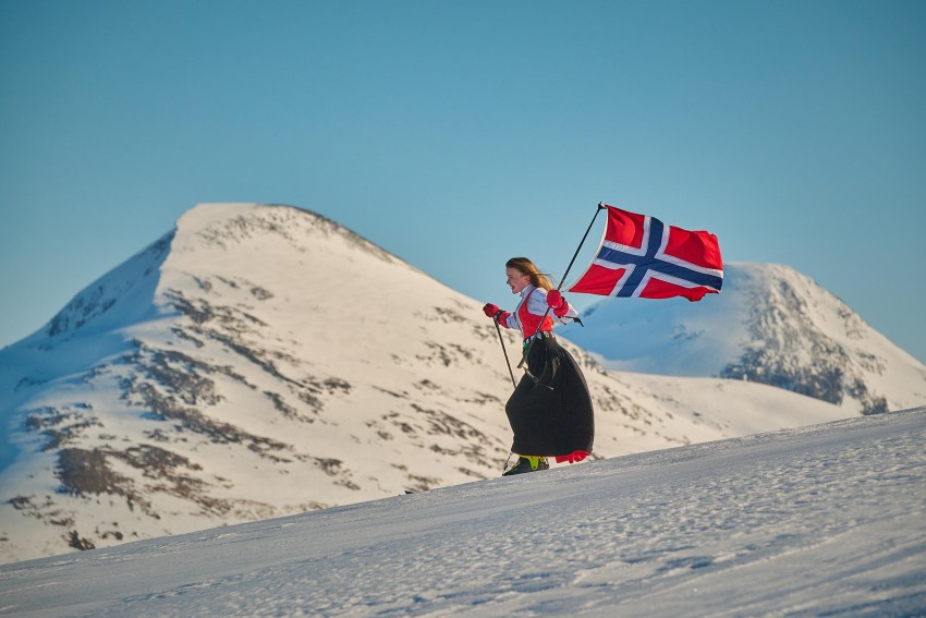 Person på ski i snødekte fjell, iført et tradisjonelt antrekk, holder et norsk flagg mot en klarblå himmel. Foto: Nathan Lediard, Lediard Foto AS.