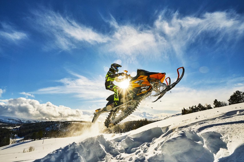 Person som kjører snøscooter opp en snødekt bakke med en strålende sol og blå himmel i bakgrunnen. Foto: Nathan Lediard, Lediard Foto AS.