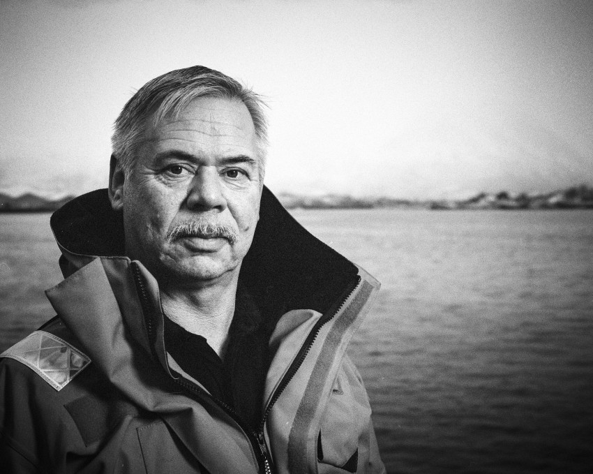 Mann i tung jakke står ved vannet med fjerne fjell. Svart-hvitt bilde. Foto: Nathan Lediard, Lediard Foto AS.