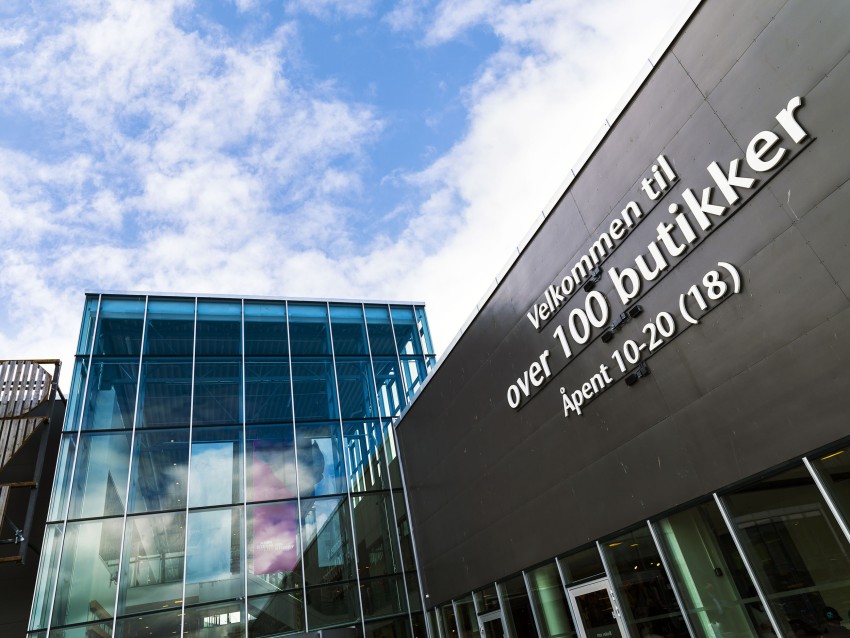 Moderne kjøpesenterinngang med glassfasade og skilting på norsk under blå himmel. Foto: Nathan Lediard, Lediard Foto AS.