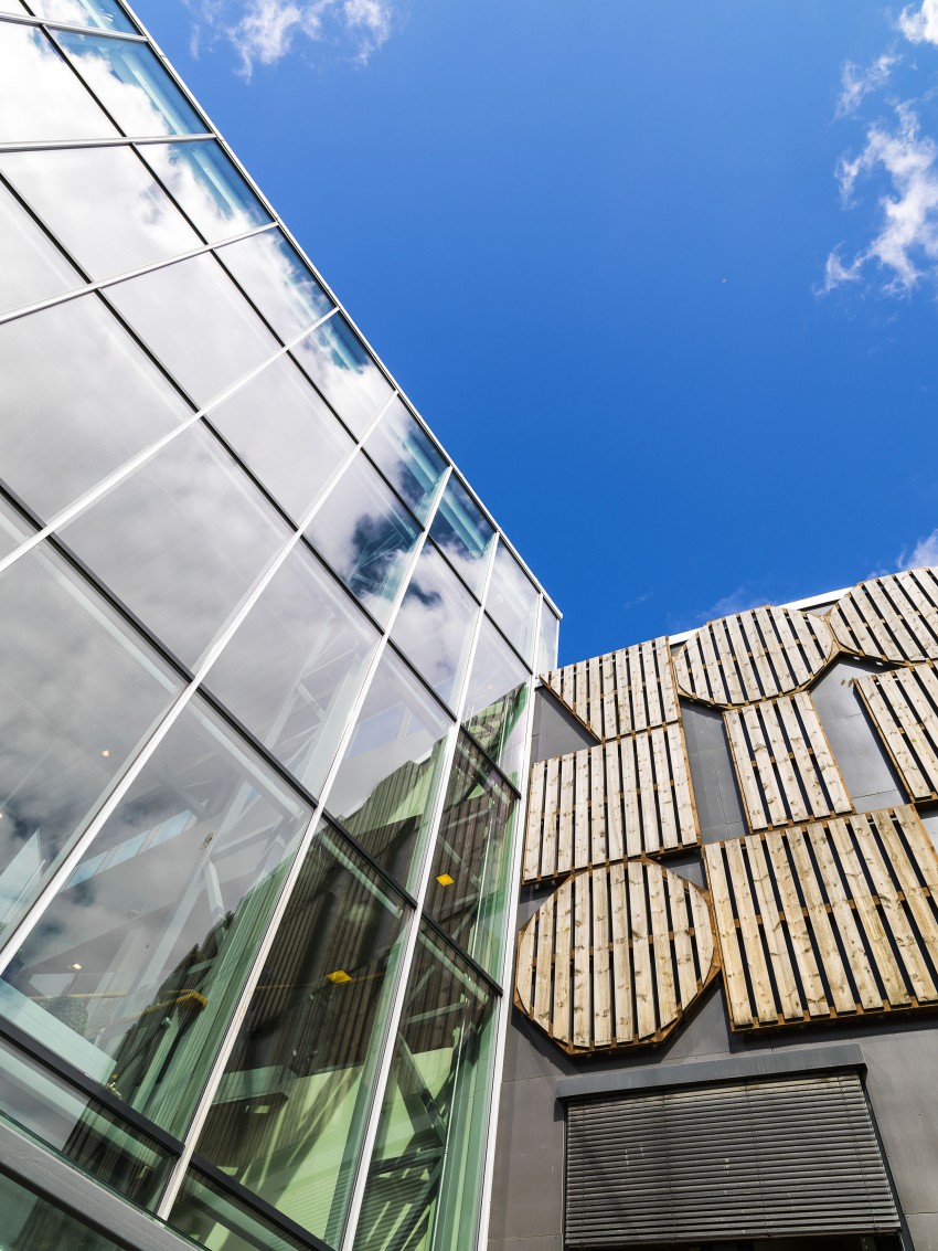 Moderne bygg med glassvinduer og trepaneler mot en knallblå himmel med skyer. Foto: Nathan Lediard, Lediard Foto AS.
