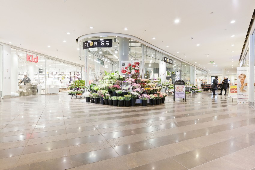 Interiør i et kjøpesenter med en blomsterbutikk som viser forskjellige fargerike blomster, og en barnebutikk til venstre. Foto: Nathan Lediard, Lediard Foto AS.