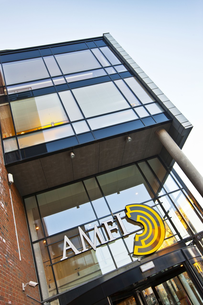 Moderne bygningsfasade med store glassvinduer, murvegg, og ordet "AMEL" i hvitt og gult på forsiden. Foto: Nathan Lediard, Lediard Foto AS.